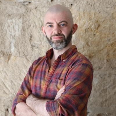 A photograph of James McEnaney standing in front of a pale stone wall. James is wearing a red, purple and orange checked shirt and stands with his arms crossed looking at the camera. He has a shaved head and a dark beard.